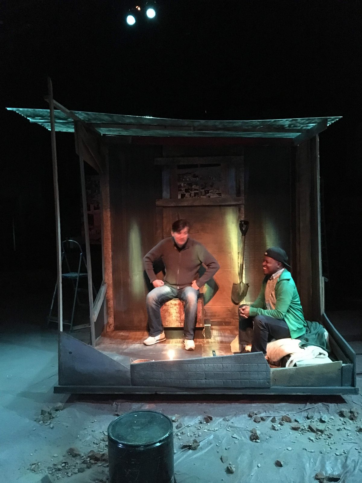 Wide view of the stage set: a corrugated shack lit warmly, with two actors seated inside